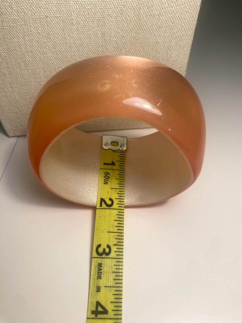 May include: A translucent orange bangle bracelet. The bracelet is measured with a yellow tape measure, showing an approximate width of 7.6 cm. The interior of the bracelet is a light beige colour.
