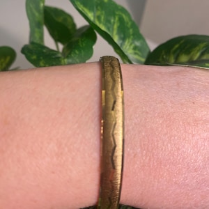 May include: A gold-colored bangle bracelet with a textured surface and a geometric design. The bracelet is worn on a wrist, with green foliage in the background. The bracelet appears to be made of metal.