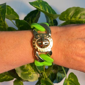 May include: A gold-tone chain bracelet with a large central clasp featuring the text "BY MARC JACOBS". The bracelet has bright green rubber links. The bracelet is worn on a wrist.