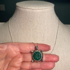 May include: A silver-toned turtle pendant necklace with a carved green stone center. The turtle pendant is held in a hand, showcasing the intricate details of the design. The necklace is displayed on a mannequin.