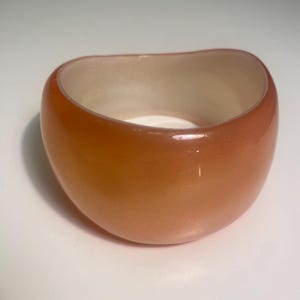 May include: A chunky, orange-toned bracelet with a rounded, oval shape. The interior is a lighter, off-white colour. The bracelet is made of a glossy material, and the overall design is simple and modern.