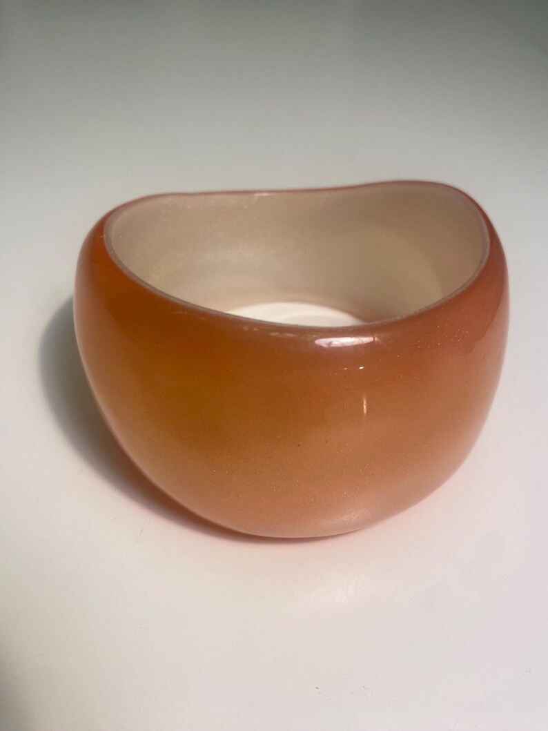 May include: A chunky, orange-toned bracelet with a rounded, oval shape. The interior is a lighter, off-white colour. The bracelet is made of a glossy material, and the overall design is simple and modern.