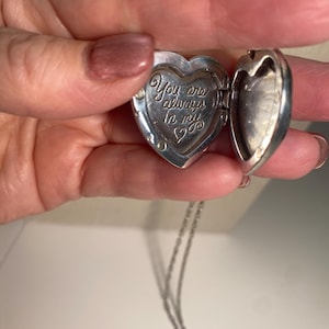 May include: Silver heart-shaped locket necklace, open to reveal the inscription "You are always in my heart." The locket is on a delicate silver chain. The locket is held open, revealing the inside.