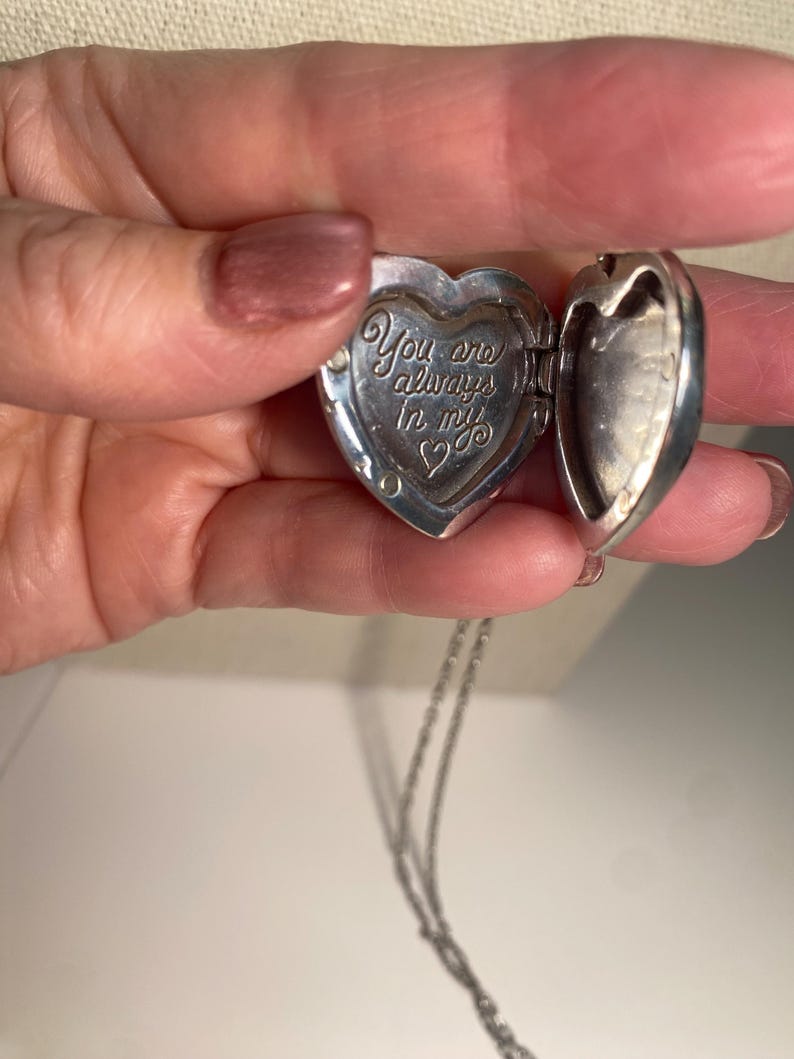 May include: Silver heart-shaped locket necklace, open to reveal the inscription "You are always in my heart." The locket is on a delicate silver chain. The locket is held open, revealing the inside.