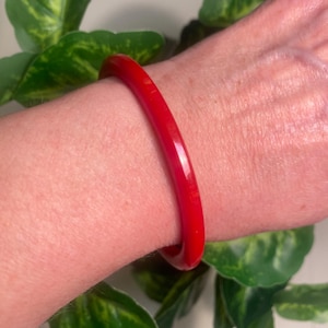 May include: A vibrant red bangle bracelet. The smooth, glossy surface of the bracelet reflects light, creating a bold and eye-catching accessory. The bracelet is a simple, circular design, perfect for everyday wear.
