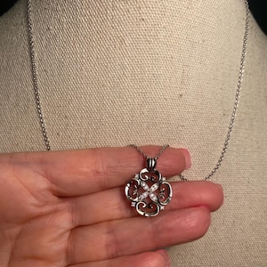 May include: Silver-toned pendant necklace featuring a four-petal floral design with clear, sparkling gemstones. The pendant hangs from a delicate chain. The necklace is displayed against a neutral background.