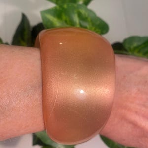 May include: A wide, peach-coloured bangle bracelet with a shimmering, glittery finish. The bracelet is a solid, curved shape and appears to be made of a smooth material. The jewellery is shown on a person's wrist.