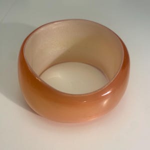 May include: A wide, orange-toned bangle bracelet. The bracelet has a smooth, glossy finish and a rounded shape. The interior of the bracelet is a lighter, peach colour. The bracelet is a solid, chunky design.