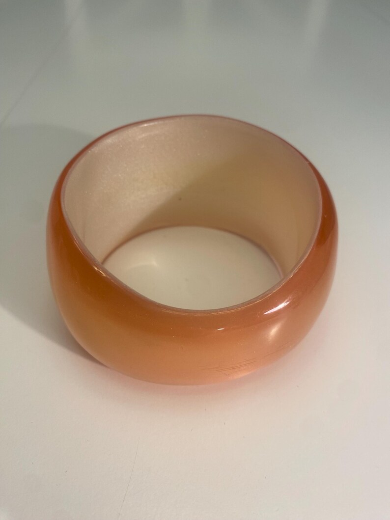 May include: A wide, orange-toned bangle bracelet. The bracelet has a smooth, glossy finish and a rounded shape. The interior of the bracelet is a lighter, peach colour. The bracelet is a solid, chunky design.