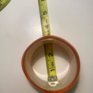 May include: An orange and cream-coloured bangle bracelet with a rounded shape. A yellow measuring tape is inside the bracelet, showing a measurement of approximately 2.5 cm. The bracelet is made of a translucent material.