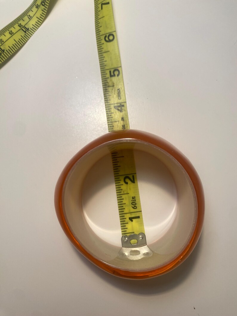 May include: An orange and cream-coloured bangle bracelet with a rounded shape. A yellow measuring tape is inside the bracelet, showing a measurement of approximately 2.5 cm. The bracelet is made of a translucent material.