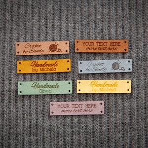 Puede incluir: Seis etiquetas de cuero de diferentes colores con texto. Las etiquetas tienen dos agujeros a cada lado. El texto en las etiquetas incluye "Crochet by Sandy", "Handmade by Michela", "Handmade Olivia", "YOUR TEXT HERE more text here".