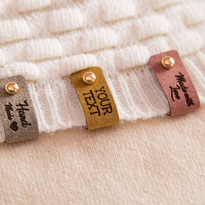 Puede incluir: Cuatro etiquetas de cuero con detalles dorados y texto diferente. Las etiquetas dicen "Hand Made", "YOUR TEXT" y "Made with Love".