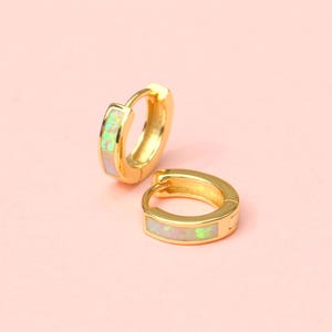 Könnte beinhalten: Zwei goldfarbene Creolen mit rechteckigem Opal-Inlay. Die Ohrringe sind vor einem zartrosa Hintergrund platziert. Der Opal hat eine Mischung aus weißen, grünen und gelben Farbtönen. Die Ohrringe sind klein und zart.