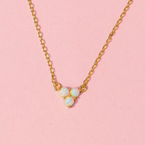Puede incluir: Un delicado collar dorado con una cadena y un colgante con tres pequeñas piedras redondas y opalescentes. El collar se muestra sobre un fondo rosa liso.