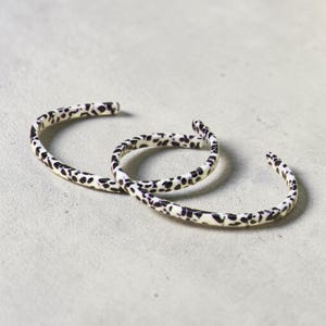 Puede incluir: Dos pulseras abiertas con un estampado de lunares en blanco y negro. Las pulseras están hechas de un material liso y tienen forma curva. Las pulseras están sobre una superficie gris claro.