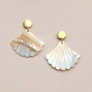 Könnte beinhalten: Ein Paar ohrringförmige Ohrringe. Jeder Ohrring hat ein fächerartiges Design mit Perlmutt-Finish, das an einem kleinen, runden, goldfarbenen Stecker befestigt ist. Die Ohrringe haben einen gewellten Rand und ein zartes, geschichtetes Aussehen.