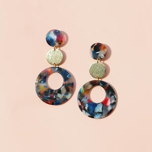 Puede incluir: Un par de pendientes colgantes coloridos. Cada pendiente presenta un colgante circular veteado con un agujero en el centro, un disco dorado y un círculo veteado más pequeño en la parte superior. Los pendientes tienen un fondo rosa.