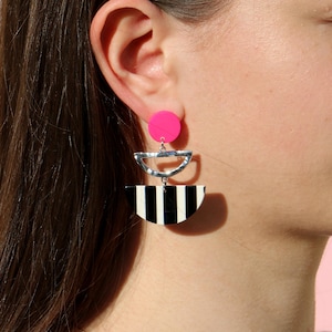 May include: A pair of dangle earrings featuring a black and white striped semi-circle, a silver semi-circle, and a hot pink circle.