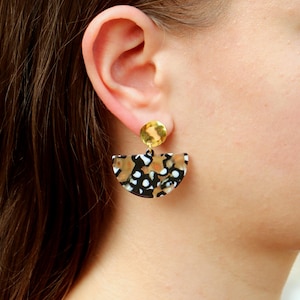 May include: A pair of gold and black statement earrings with a semi-circle shaped design. The earrings feature a black and white patterned design with a gold accent.