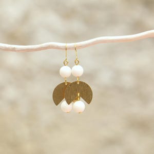 May include: Gold earrings with white beads and a geometric design. The earrings are hanging from a white branch.