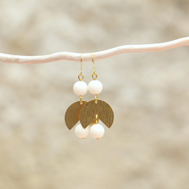 May include: Gold earrings with white beads and a geometric design. The earrings are hanging from a white branch.