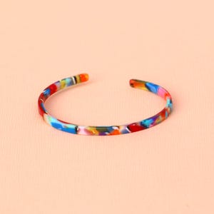 Könnte beinhalten: Ein mehrfarbiges offenes Armband mit einem Muster aus Rot, Blau, Gelb und Grün. Das Armband besteht aus glänzendem Material und hat eine glatte, abgerundete Form. Das Armband wird auf einem hellpfirsichfarbenen Hintergrund gezeigt.