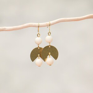 May include: Gold earrings with white beads and a geometric design. The earrings are hanging from a white branch.