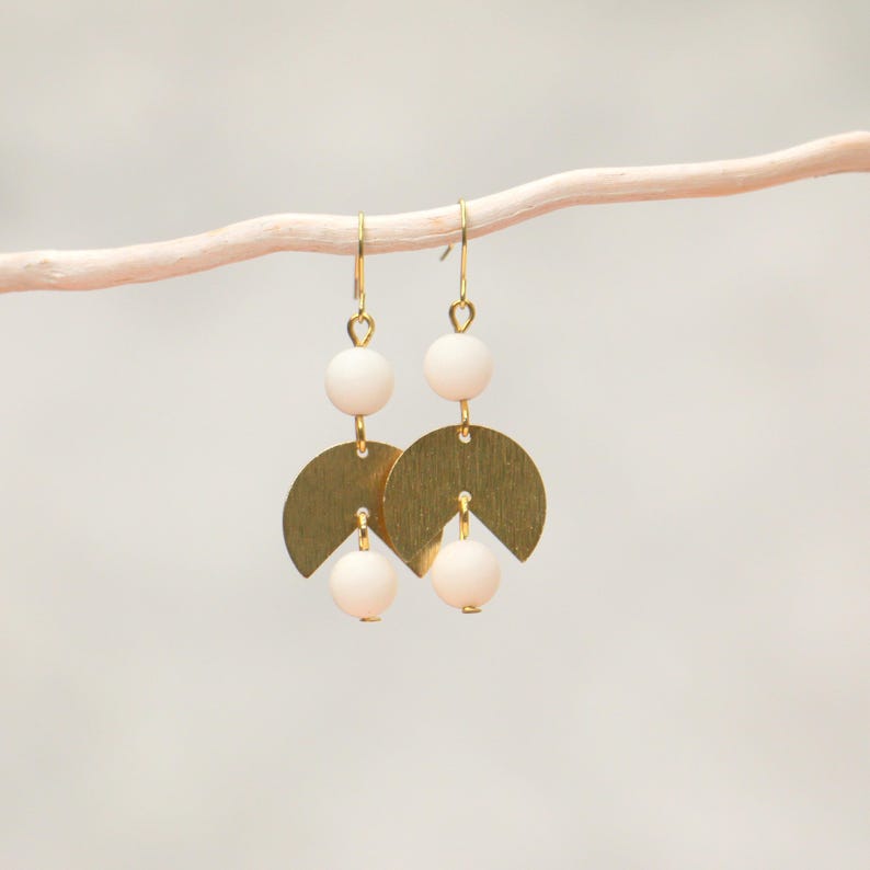 May include: Gold earrings with white beads and a geometric design. The earrings are hanging from a white branch.