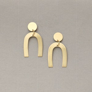 Peut inclure: Une paire de boucles d'oreilles dorées au design simple et moderne. Chaque boucle d'oreille présente une pièce incurvée en forme de U avec un petit disque rond au sommet.