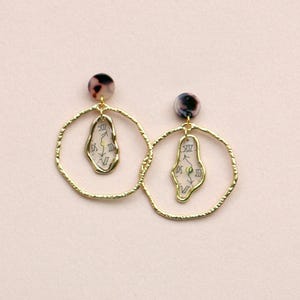 Pode incluir: Um par de brincos de argola dourados com acabamento texturizado. Cada brinco apresenta um pequeno design de relógio abstrato dentro de uma forma de lágrima, suspenso dentro do aro. A parte superior de cada brinco tem um pequeno pino redondo com padrão de tartaruga.