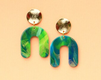May include: A pair of earrings with a gold-colored circular stud and a marbled, U-shaped pendant. The pendant features shades of green and blue. The earrings hang against a peach-colored background.