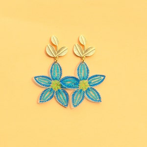 Puede incluir: Un par de pendientes florales azules y verdes con detalles de hojas doradas. Los pendientes están hechos de acrílico transparente y tienen un diseño delicado y femenino.