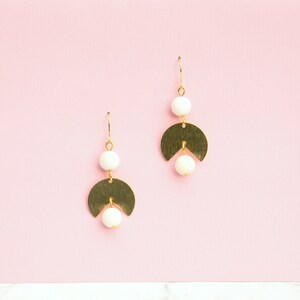 May include: A pair of gold earrings with white beads and a crescent moon shape. The earrings are on a pink background.
