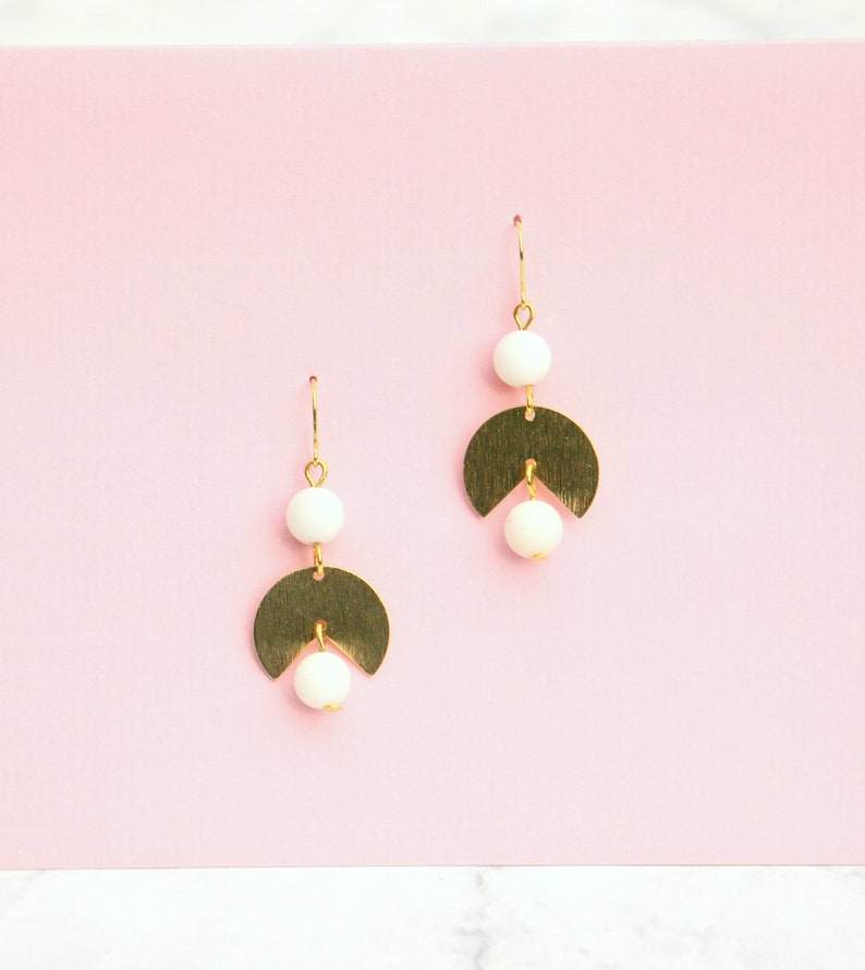 May include: A pair of gold earrings with white beads and a crescent moon shape. The earrings are on a pink background.