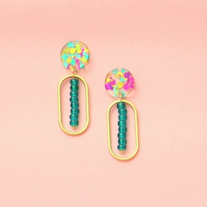 Könnte beinhalten: Ein Paar goldene Ohrringe mit einem Design aus blaugrünen Perlen. Die Ohrringe haben einen klaren, runden oberen Teil mit buntem Konfetti darin.