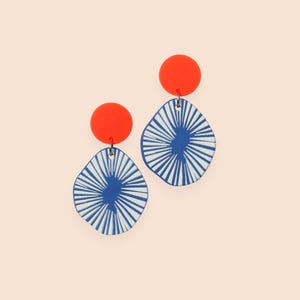 Könnte beinhalten: Ein Paar Ohrringe mit einem auffälligen Design. Jeder Ohrring hat einen leuchtend orangefarbenen runden Stecker, der mit einem blau-weiß gemusterten Anhänger verbunden ist. Der Anhänger hat eine einzigartige Form mit radialen Linien, die einen auffälligen visuellen Effekt erzeugen.
