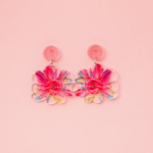 Może przedstawiać: Kolczyki w kwiaty na różowym tle. Kolczyki mają przezroczysty kształt kwiatu i zdobione są żywym różowo-czerwonym wzorem kwiatowym. Każdy kolczyk ma mały, okrągły, różowy kołek.