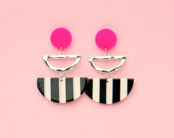 May include: A pair of statement earrings with a black and white striped half-circle design, a silver half-circle, and a hot pink circle at the top.