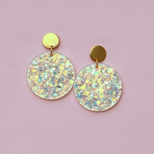 Pode incluir: Um par de brincos de disco iridescentes cheios de glitter. Cada brinco apresenta um pino circular dourado e um pingente redondo maior preenchido com glitter brilhante multicolorido. Em um fundo rosa suave.