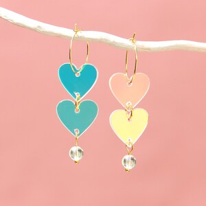 May include: Two pairs of heart-shaped earrings. One pair features turquoise and teal hearts, the other has peach and yellow hearts. Each heart is connected with a gold hoop and has a clear bead at the bottom. The background is a solid pink.