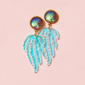 Puede incluir: Un par de pendientes con tachuelas circulares doradas con escamas iridiscentes. Debajo, un diseño en forma de medusa azul claro con acentos blancos cuelga de la tachuela. Los pendientes están colocados sobre un fondo rosa suave.