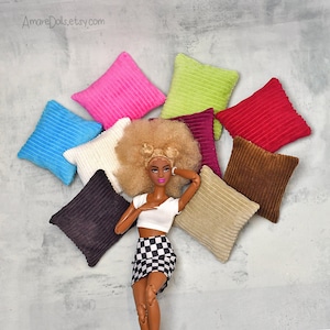 Puede incluir: Una muñeca con el pelo rubio y una falda a cuadros está rodeada de ocho almohadas cuadradas de varios colores, incluyendo rosa, verde, rojo, marrón, azul, blanco y beige. Las almohadas tienen una textura de pana.