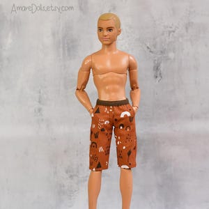 May include: A doll wearing brown shorts with a white and black abstract pattern. The doll has short blonde hair and is shirtless. The shorts have a dark brown waistband and pockets. The doll is standing in front of a gray background.