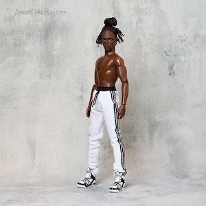 May include: A black doll with a brown complexion wearing white sweatpants with black stripes down the sides and black and white trainers. The doll has a bun in their hair and is standing against a grey background.