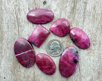 May include: A selection of polished gemstones in shades of magenta, with oval and irregular shapes. The stones have darker and lighter veining. A coin is included for size reference. The gemstones are displayed on a weathered wooden surface.