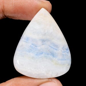 May include: A pear-shaped gemstone with a polished surface. The stone features a white base with wavy blue and light yellow patterns. The gemstone is held between two fingers against a black background.