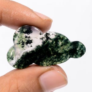 Pode incluir: Uma pedra preciosa em forma de nuvem com um padrão de ágata musgo. A pedra apresenta uma mistura de cores brancas, verdes e pretas. A pedra preciosa é segurada entre dois dedos.