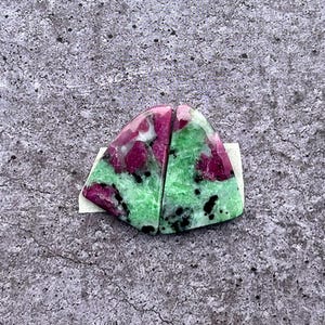 Op de afbeelding: Twee gepolijste zoisietstenen met robijninsluitsels. De stenen zijn een mix van groen, roze en zwart, met een driehoekige vorm. De robijnsecties zijn diep roze, terwijl de zoisiet levendig groen is.
