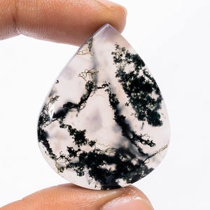 Pode incluir: Uma pedra preciosa de ágata musgo em forma de gota. A pedra translúcida apresenta inclusões dendríticas pretas e verdes, que se assemelham a padrões vegetais, sobre um fundo pálido. A pedra é segurada entre dois dedos.